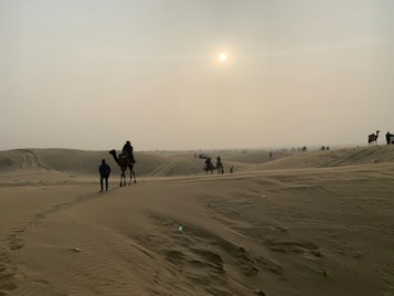 A vast desert landscape with rolling sand dunes under a hazy sky. Several camels, some with riders, are dispersed across the scene, with the sun partially obscured by thin clouds, casting a warm glow. The scene includes a few people on foot and the terrain is mostly barren.