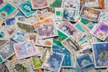 A diverse collection of vintage postage stamps with various designs, each featuring different colors, patterns, and illustrations. Some stamps depict portraits of people, architectural landmarks, and cultural symbols. The layout is disorganized, with stamps overlapping one another, creating a colorful and intricate mosaic.