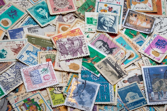 A diverse collection of vintage postage stamps with various designs, each featuring different colors, patterns, and illustrations. Some stamps depict portraits of people, architectural landmarks, and cultural symbols. The layout is disorganized, with stamps overlapping one another, creating a colorful and intricate mosaic.