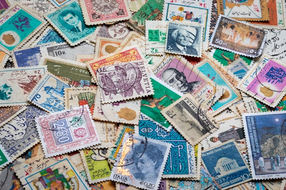 A diverse collection of vintage postage stamps with various designs, each featuring different colors, patterns, and illustrations. Some stamps depict portraits of people, architectural landmarks, and cultural symbols. The layout is disorganized, with stamps overlapping one another, creating a colorful and intricate mosaic.