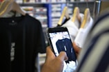 A person is using a smartphone to take a photo of a black t-shirt hanging on a rack. The shirt has some text or a logo on it. There are other clothes on hangers next to it, and a shelf with various items is visible in the background.