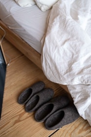 A neatly made bed with white bedding and wooden frame is seen, with a pair of grey felt slippers placed beside it on a wooden floor.