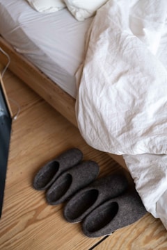 A neatly made bed with white bedding and wooden frame is seen, with a pair of grey felt slippers placed beside it on a wooden floor.