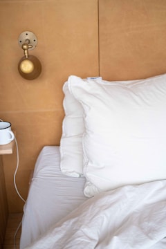 A neatly made bed with a white pillow and duvet. A small bedside shelf holds a cup, and a brass wall-mounted lamp is positioned above. The backdrop is a light brown wooden or faux leather panel.