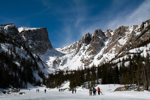 A group of people are walking across a snow-covered area surrounded by tall pine trees and majestic, rocky mountains. The peaks are capped with snow, and the sky is a clear, deep blue.