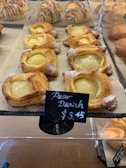 Several golden-brown pastries filled with a creamy pear mixture are displayed on a tray. A small black sign indicates they are Pear Danish, priced at $3.45. The pastries are surrounded by other baked goods, including croissants in the background.