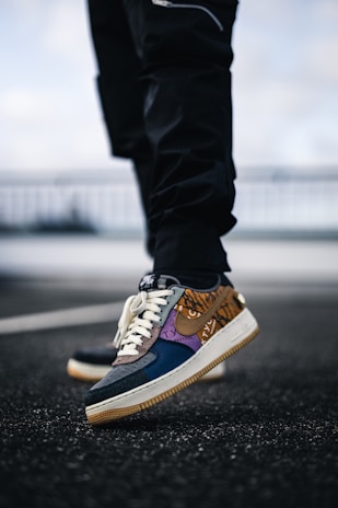 A close-up of a colorful sneaker with a unique design, featuring a mix of patterns and textures. The sole is brown and the shoe is being worn by someone in black pants, with the background blurred.