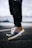 A close-up of a colorful sneaker with a unique design, featuring a mix of patterns and textures. The sole is brown and the shoe is being worn by someone in black pants, with the background blurred.
