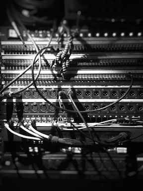 A black and white close-up of a complex audio equipment setup with numerous cables and connectors. The scene includes various knobs, input jacks, and twisted cords, with some light and shadow creating a high contrast effect.