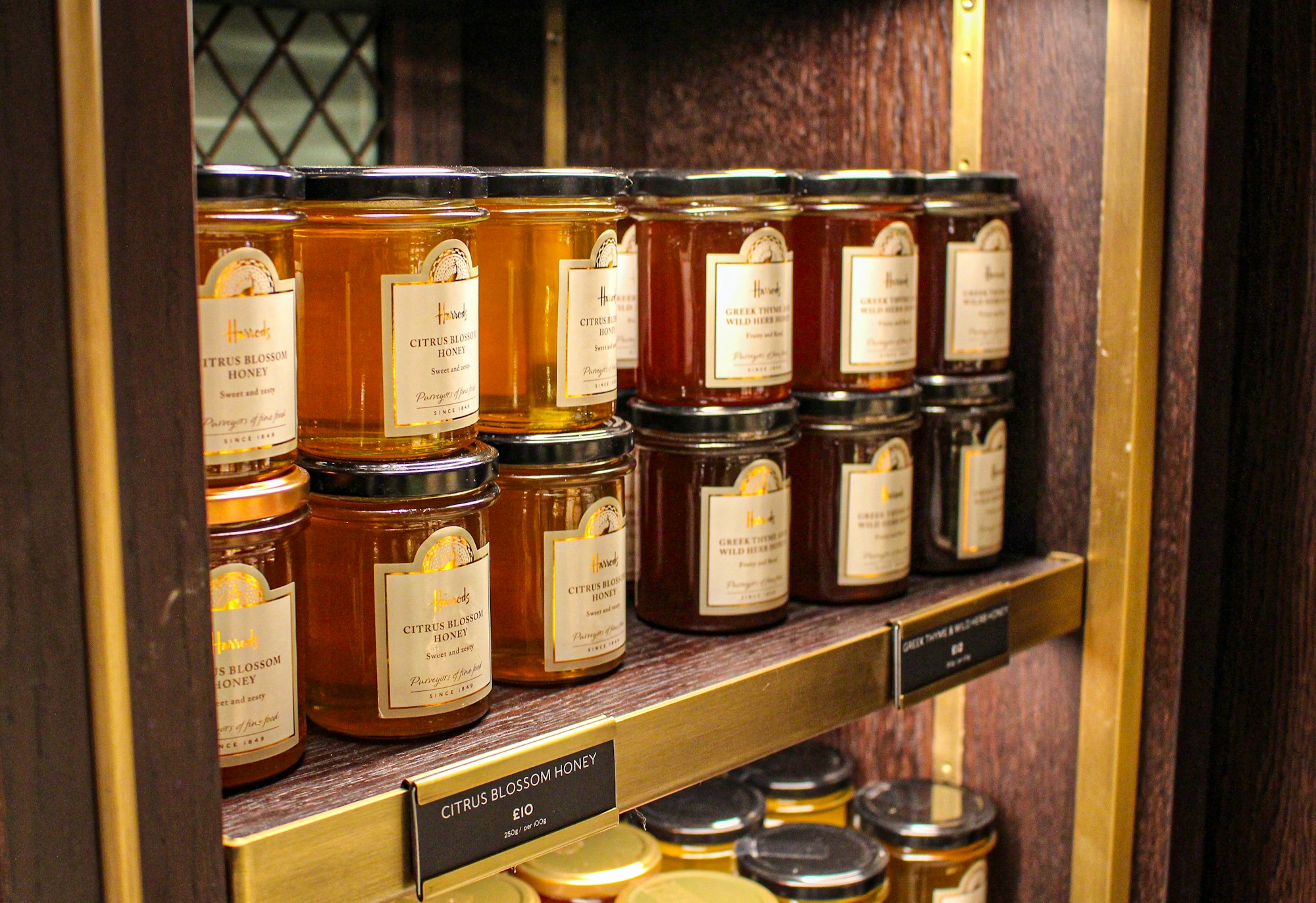 Honey, jams, preserves, marmalades in Harrods, Brompton Road, London, Great Britain: Citrus Blossom Honey, Greek Thyme Wild Herbs Honey, all in jars on wooden shelves.