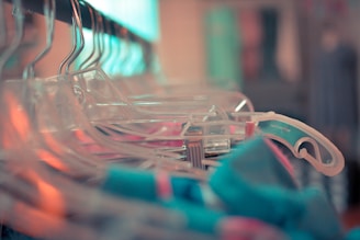 A close-up view of several transparent plastic hangers arranged on a clothing rack, with some colorful garments visible. The focus is on the hangers, creating a shallow depth of field effect with a blurred background.
