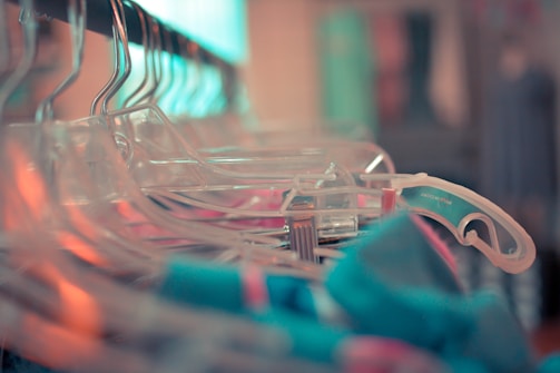 A close-up view of several transparent plastic hangers arranged on a clothing rack, with some colorful garments visible. The focus is on the hangers, creating a shallow depth of field effect with a blurred background.