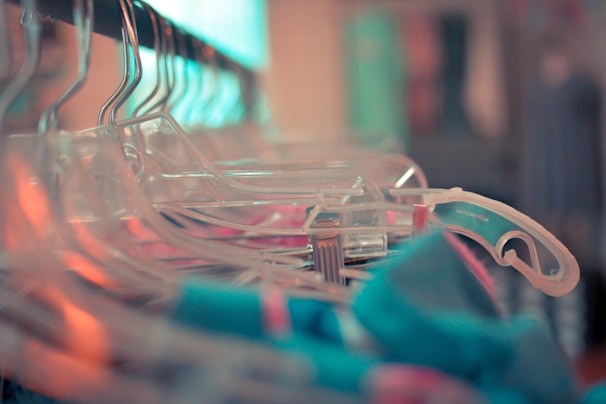 A close-up view of several transparent plastic hangers arranged on a clothing rack, with some colorful garments visible. The focus is on the hangers, creating a shallow depth of field effect with a blurred background.
