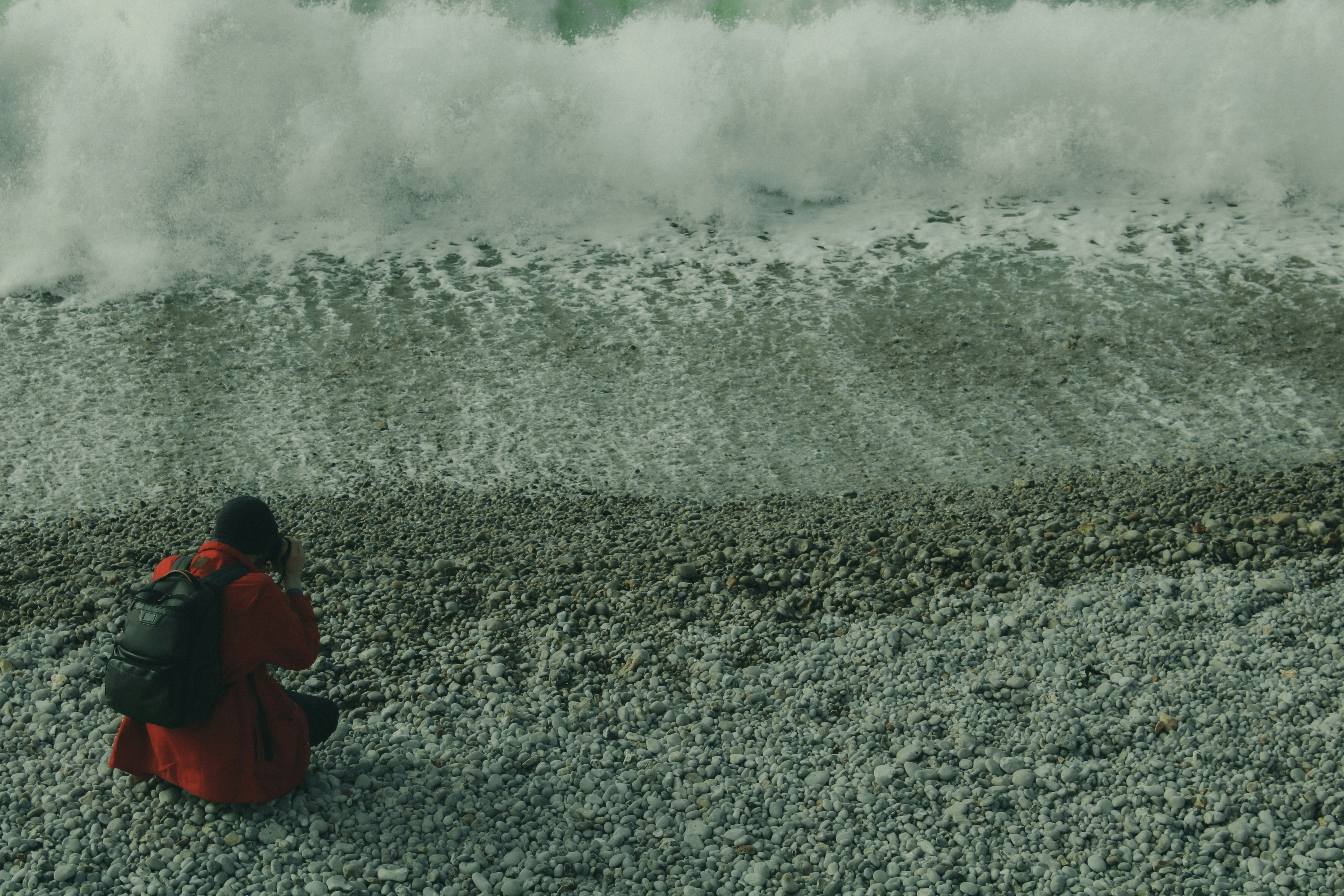 Persona con sudadera con capucha roja sentada en arena gris cerca de las olas del océano durante el día