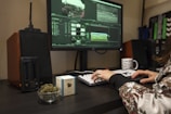 A person is using a desktop computer for video editing, visible through a software interface on the screen. A cup of coffee, a small bowl of cannabis buds, and a box labeled with a cannabis brand are on the desk. Audio equipment and a modern vaporizer are also present.