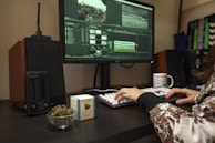 A person is using a desktop computer for video editing, visible through a software interface on the screen. A cup of coffee, a small bowl of cannabis buds, and a box labeled with a cannabis brand are on the desk. Audio equipment and a modern vaporizer are also present.