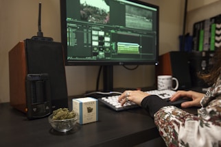 A person is using a desktop computer for video editing, visible through a software interface on the screen. A cup of coffee, a small bowl of cannabis buds, and a box labeled with a cannabis brand are on the desk. Audio equipment and a modern vaporizer are also present.