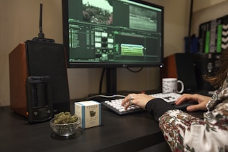 A person is using a desktop computer for video editing, visible through a software interface on the screen. A cup of coffee, a small bowl of cannabis buds, and a box labeled with a cannabis brand are on the desk. Audio equipment and a modern vaporizer are also present.