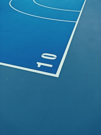 A section of a sports court with a vibrant blue surface. The image shows the corner area with a white boundary line and the number '10' marked prominently. The curvature of additional painted lines can be seen in the upper part of the image, indicating the layout of the playing area.