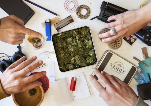 Various items are scattered on a table, including a metal tin filled with cannabis, a red lighter, and rolling papers. Two hands are interacting with the items, one holding some rolling paper. Other objects include a grinder, headphones, and a smartphone.