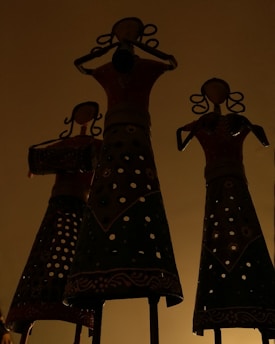 Three intricately designed metal sculptures of women, featuring delicate patterns and ornate hairstyles, stand prominently against a muted background. Their attire is adorned with fine geometric and floral motifs, creating a play of light and shadow.