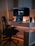 A modern workspace setup featuring a large curved monitor and a laptop on a sleek white desk. A black mesh office chair is positioned in front of the desk. Ambient lighting casts a warm glow, enhancing the room's cozy atmosphere. Decorative items include a small statue and potted plant, while camera lenses and a smartphone are neatly arranged nearby.