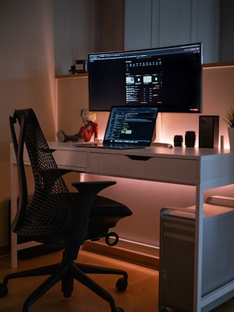 A modern workspace setup featuring a large curved monitor and a laptop on a sleek white desk. A black mesh office chair is positioned in front of the desk. Ambient lighting casts a warm glow, enhancing the room's cozy atmosphere. Decorative items include a small statue and potted plant, while camera lenses and a smartphone are neatly arranged nearby.