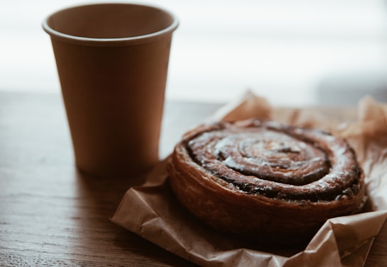 A warm cinnamon roll sits wrapped in brown parchment paper on a wooden surface, next to a plain brown disposable coffee cup. The background is softly blurred, giving a cozy and inviting feel.