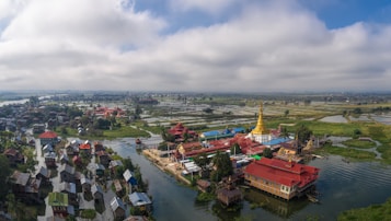 A scenic landscape with numerous stilt houses atop a lake, lush green vegetation, and an ornate golden pagoda in the center. The area is surrounded by water channels and fields stretching into the horizon under a partly cloudy sky.