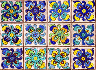 A collection of colorful decorative tiles featuring intricate floral patterns. Each tile displays a symmetrical design with vivid hues of blue, green, yellow, and red, arranged in a repetitive grid.
