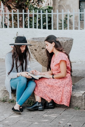 Two women are sitting outdoors next to a concrete structure, engage in reading a book together. One woman wears a black hat, blue jeans, and a long-sleeve sweater, while the other wears a pink floral dress and black shoes. A white metal fence and some greenery can be seen in the background.