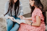 Two women are seated outdoors and looking at an open book, seemingly engaged in discussion or study. One woman is wearing a striped shirt with a grey cardigan and jeans, while the other is dressed in a pink floral dress. They appear focused and interested in the content of the book.