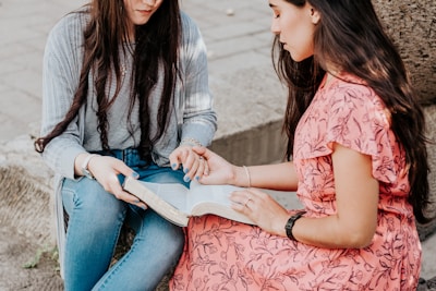 Two women are seated outdoors and looking at an open book, seemingly engaged in discussion or study. One woman is wearing a striped shirt with a grey cardigan and jeans, while the other is dressed in a pink floral dress. They appear focused and interested in the content of the book.