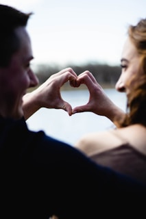 A close-up of two people facing each other, forming a heart shape with their hands. The background appears to be a blurred natural setting, possibly near a body of water.