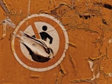 A weathered and peeling safety sign against a textured, rusty orange wall. The sign features a figure inside a prohibition circle, partially obscured by peeling layers of the sign itself.