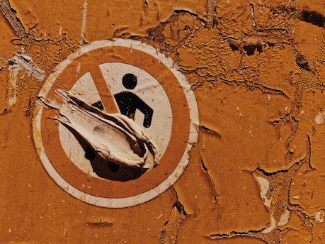 A weathered and peeling safety sign against a textured, rusty orange wall. The sign features a figure inside a prohibition circle, partially obscured by peeling layers of the sign itself.