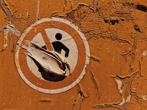 A weathered and peeling safety sign against a textured, rusty orange wall. The sign features a figure inside a prohibition circle, partially obscured by peeling layers of the sign itself.