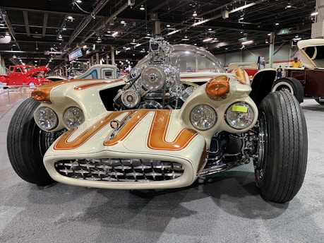 A custom, futuristic car showcased indoors with a prominent display of its intricate engine and unique design. The car features a white body with brown accents, multiple headlamps, and a wide, aggressive front grille. It is set against the backdrop of a car show, with various other classic and vintage vehicles visible in the background under bright, artificial lighting.