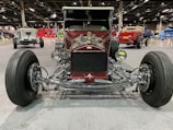 A vintage hot rod car is prominently displayed in an indoor exhibition hall. The vehicle has a deep burgundy color with chrome accents and large, exposed wheels. Other vintage cars in various colors are visible in the background, enhancing the automotive theme of the setting.