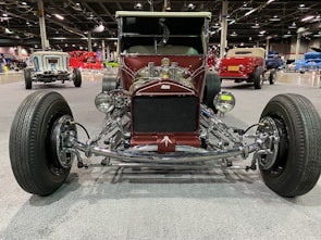 A vintage hot rod car is prominently displayed in an indoor exhibition hall. The vehicle has a deep burgundy color with chrome accents and large, exposed wheels. Other vintage cars in various colors are visible in the background, enhancing the automotive theme of the setting.