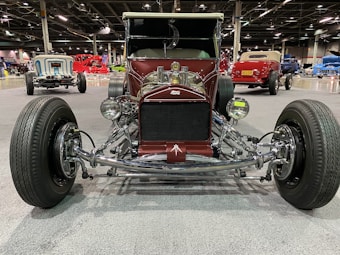 A vintage hot rod car is prominently displayed in an indoor exhibition hall. The vehicle has a deep burgundy color with chrome accents and large, exposed wheels. Other vintage cars in various colors are visible in the background, enhancing the automotive theme of the setting.