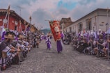 A vibrant procession occurs on a cobblestone street, featuring people dressed in historical or religious attire, likely for a cultural or religious event. Participants hold spears and wear elaborate garments, with some in purple robes and others in Roman-style armor. The procession is flanked by colorful colonial buildings with a mountainous backdrop under a cloudy sky.