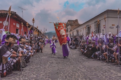 A vibrant procession occurs on a cobblestone street, featuring people dressed in historical or religious attire, likely for a cultural or religious event. Participants hold spears and wear elaborate garments, with some in purple robes and others in Roman-style armor. The procession is flanked by colorful colonial buildings with a mountainous backdrop under a cloudy sky.