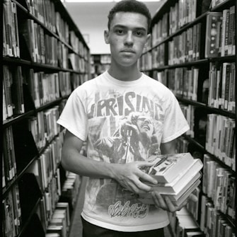 A young person stands in a library aisle, holding several books. The individual is wearing a graphic t-shirt featuring an image and text. Books are neatly arranged on shelves that line the aisle, creating a sense of depth.