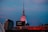 A cityscape at dusk featuring the Museum of Science, with its illuminated sign on a building. The sky has a gradient of blue and pink hues, while some of the buildings in the surroundings are also lit up. A tall tower with a distinctive spire is prominently visible.