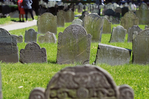 A series of aged, weathered gravestones are placed in a well-maintained graveyard with vibrant green grass. The stones display intricate carvings and inscriptions. In the background, there is a walkway with two people standing near the graves.