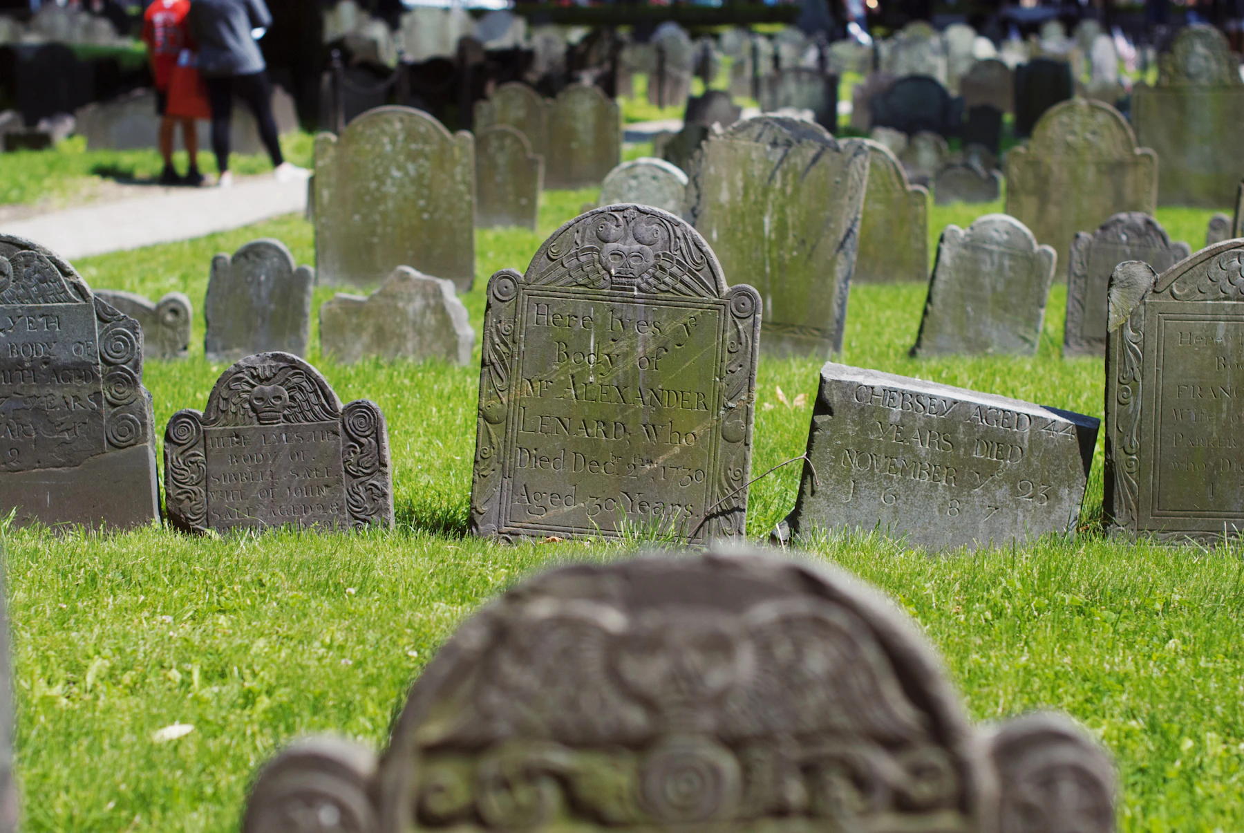 Rows of dark slate headstones with carved arched tops in an old burying ground, two visitors walking the path behind them