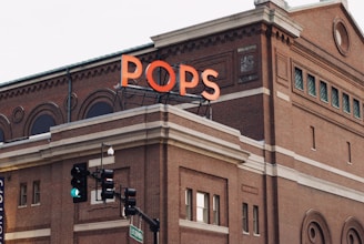 A brick building with decorative architectural features, including arched and rectangular windows. The sign 'POPS' is prominently displayed in large, bright orange letters on the roof. Below, a traffic light is visible showing a green signal, and a street sign labeled St. Spring is nearby.