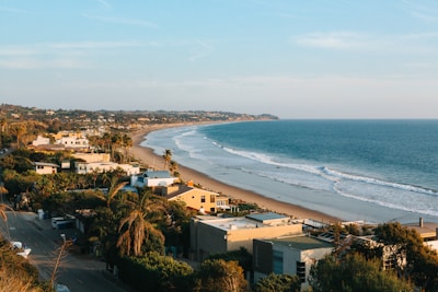 A coastal scene featuring a stretch of sandy beach lined with oceanfront houses and palm trees. The ocean is calm with gentle waves reaching the shore. The background includes rolling hills covered in greenery, and the sky is clear with a few wispy clouds.