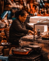 An individual sits at a food stall, preparing and arranging various dishes. The scene includes cooking utensils, trays of food items like spring rolls, and containers. The setting suggests a busy marketplace with a variety of items visible in the background.